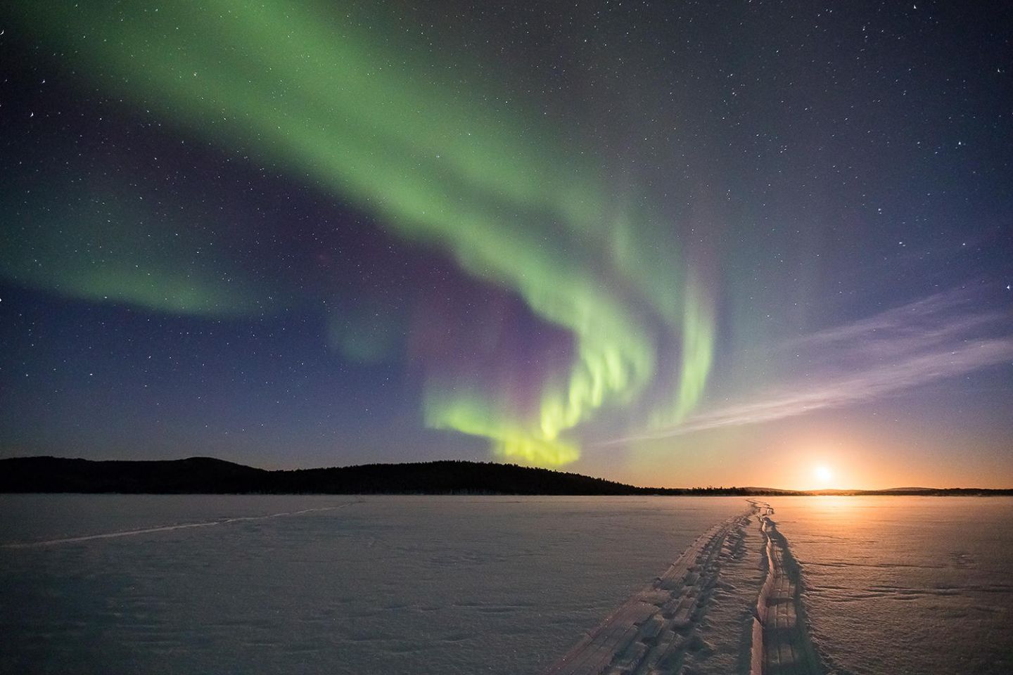 Snowmobile tracks on a frozen lake leading to the rising moon under the Aurora Borealis in northern Finnish Lapland