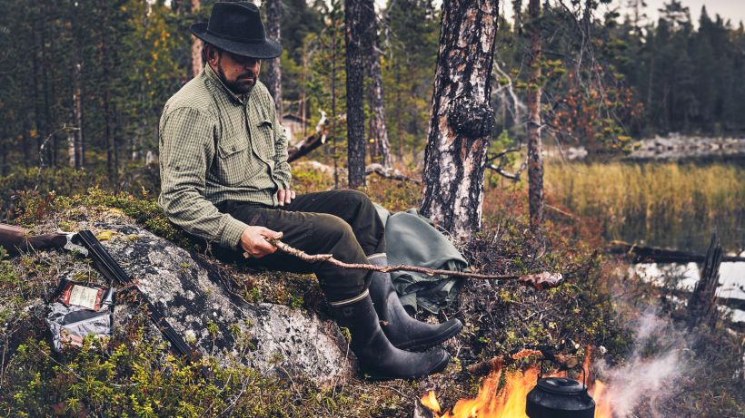 Mies istuu mättäällä ja paistaa makkaraa nuotiolla.